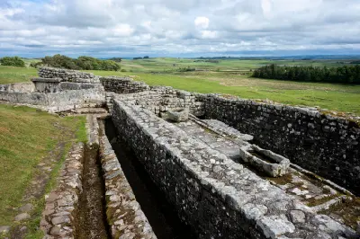 Ancient stone walls and archaeological remains at Housesteads Roman Fort beside Hadrian's Wall, with green Northumberland hills beneath a cloudy sky.