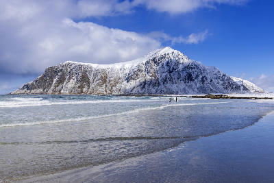 Snow-covered mountains rising above Unstad Beach on Vestvågøya in the Lofoten Islands, with winter waves along the sandy shoreline in Nordland, Norway.