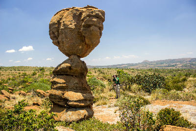 A large natural balancing rock formation stands among rocky bushveld vegetation in the Magaliesberg Mountains, with rolling hills and open countryside visible in the distance, North West Province, South Africa.