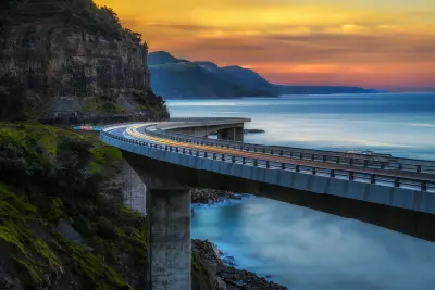 Long-exposure view of Sea Cliff Bridge at sunset, with vehicle light trails curving above the Pacific Ocean beneath a glowing orange sky.
