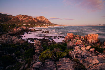 Rocky shoreline and ocean waves at sunset along the Hermanus Cliff Path with Walker Bay and mountain backdrops in South Africa.