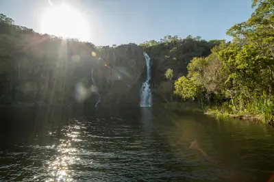 Two waterfalls cascading down rocky cliffs into a broad plunge pool, with bright sunlight, lens flare and reflections on the water at Wangi Falls, Litchfield National Park, Northern Territory, Australia.