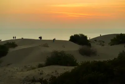 Golden sunset over the Mediterranean Sea and rolling sand dunes at Patara, with scattered shrubs and visitors silhouetted along the dune ridges.