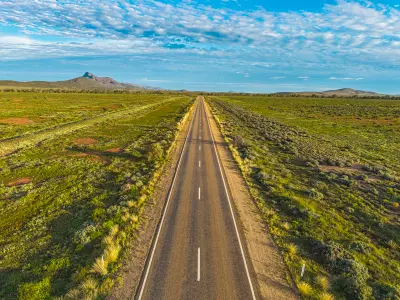 Straight sealed Outback Highway crossing green plains near Mern Merna Station, with Mount Aleck and distant Flinders Ranges mountains beneath a blue, cloud-filled sky.