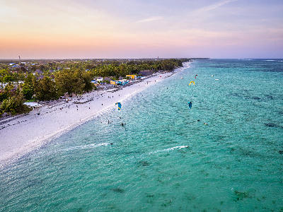 Drone view of kiteboarders on the turquoise waters of Paje Beach with white sand, palm trees, and the Zanzibar coastline at sunset.