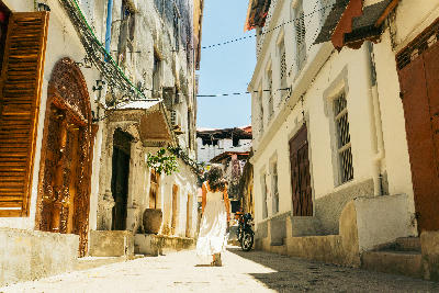Woman in a white dress walking along a narrow alley lined with carved doors and historic buildings in Stone Town, Zanzibar.