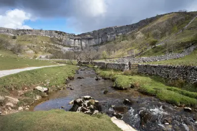 Malham Cove limestone cliffs above a rocky stream, grassy valley and traditional dry-stone walls beneath partly cloudy skies in Yorkshire Dales National Park.
