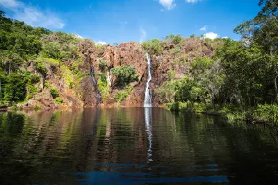 Wide view of two waterfalls cascading down rocky cliffs into a large reflective plunge pool surrounded by green tropical woodland at Wangi Falls, Litchfield National Park, Northern Territory, Australia.