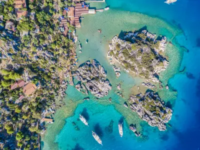 Top-down aerial view of Kaleköy and the Simena area, with rocky islets surrounded by crystal-clear turquoise water, small boats anchored offshore and buildings tucked along the rugged coast.