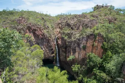 Wide view of Tolmer Falls cascading down a tall sandstone cliff into a dark plunge pool, with rocky escarpments and green woodland in Litchfield National Park, Northern Territory, Australia.