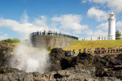 Seawater erupts from the Kiama Blowhole as visitors watch from viewing platforms beneath the white Kiama Lighthouse.