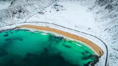 Drone view of Haukland Beach on Vestvågøya in winter, with turquoise Arctic water, golden sand, a curving coastal road, and snow-covered mountains in the Lofoten Islands, Norway.