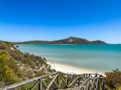 Wooden boardwalk descending to a white-sand beach at Kraalbaai, with turquoise lagoon waters and the Postberg Peninsula in the background.