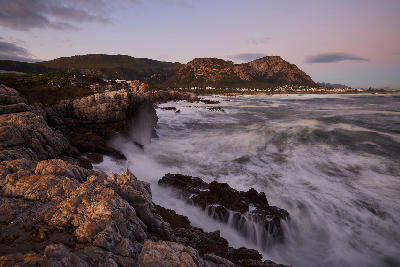 Sunset surf and rocky coastline along Hermanus Cliff Path with Walker Bay mountains in the Western Cape of South Africa.