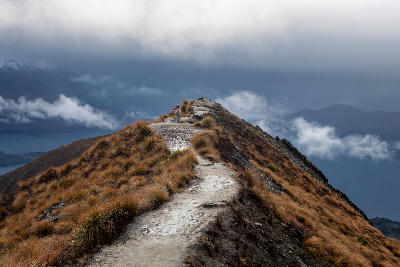 Hiker on the ridge path near Roys Peak summit overlooking Lake Wanaka and mountains in Wanaka, South Island, New Zealand.