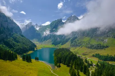 Elevated view of Seealpsee surrounded by green alpine slopes, forest, and low cloud drifting across mountain ridges in the Alpstein mountains, Switzerland.