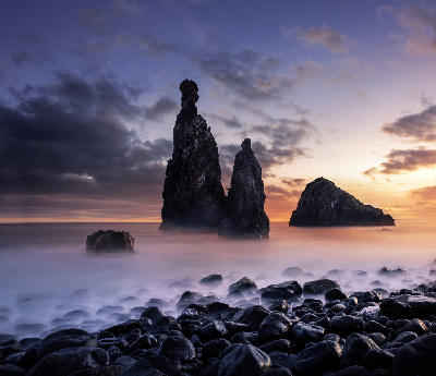 Sunrise over the Ilhéu da Ribeira da Janela rock formations, with smooth long-exposure waves and dark volcanic pebbles in the foreground on Madeira Island, Portugal.