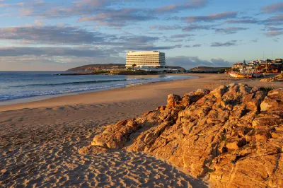 Central Beach in Plettenberg Bay looking towards Beacon Isle, with sandy shoreline, rocky foreground and calm ocean waves at sunrise.