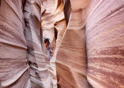 Photographer with a tripod between narrow pink and red striped sandstone walls in Zebra Slot Canyon, Utah.
