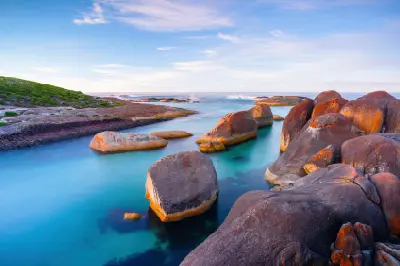 Large orange-tinted granite boulders sitting in a calm, vibrant turquoise channel along a rocky coastline under a pastel blue sky at dusk.