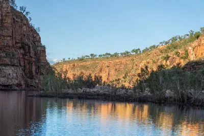 Calm water reflects the sunlit red sandstone walls and riverside vegetation of Chamberlain Gorge.