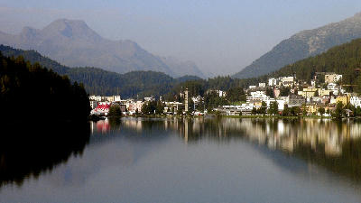 Lake St. Moritz at dawn with town buildings and mountains reflected in calm water, Engadin, Switzerland.