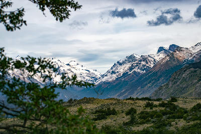 Mountain landscape seen from Pliegue Tumbado trail with foreground vegetation and snow-capped peaks under cloudy skies in Patagonia, Argentina.
