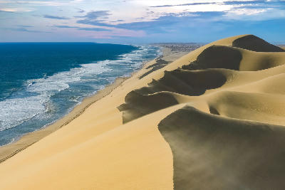 Towering golden sand dunes descending steeply to the Atlantic shoreline along Namibia’s Skeleton Coast, with rolling ocean waves stretching into the distance beneath a partly cloudy sky.