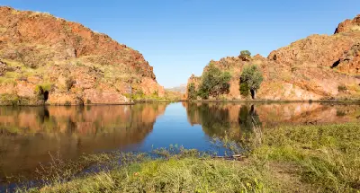 Calm water reflects rugged orange rock walls and native vegetation beside Lake Argyle in the Kimberley.