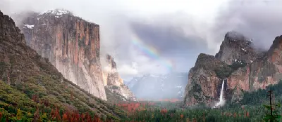 Tunnel View Yosemite Valley California with El Capitan, Bridalveil Fall and rainbow over forested valley under cloudy sky.