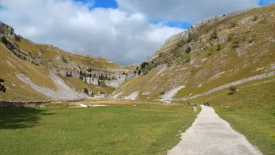 Gravel walking path winding between grassy limestone slopes and rocky cliffs at Gordale Scar, North Yorkshire, England.