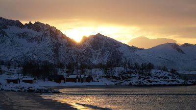 Red cabins on Bøvær beach at sunset with snow-covered mountains and calm coastal water on Senja island, Norway