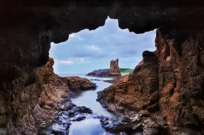 View through a dark coastal rock cave toward Cathedral Rocks, with rocky channels and calm water leading toward the formation.