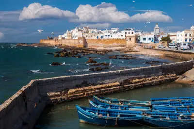 Blue fishing boats sit beneath the fortified harbour walls of Essaouira on Morocco’s Atlantic coast, with whitewashed medina buildings and rocky shoreline extending along the ocean.