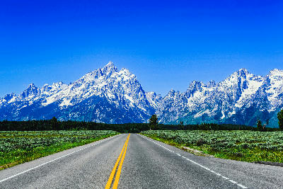 Empty road with yellow center line leading toward the snow-capped Grand Teton Range under a clear blue sky in Wyoming.