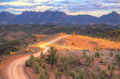 Winding dirt road over a sunlit ochre ridge with rugged Flinders Ranges mountains in the background, South Australia.