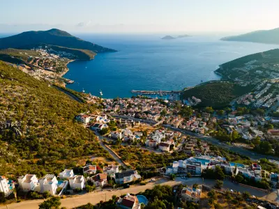 Panoramic aerial view of Kalkan, with white hillside villas and boutique hotels descending toward the marina and deep-blue Mediterranean Sea, framed by rugged green headlands and distant islands.