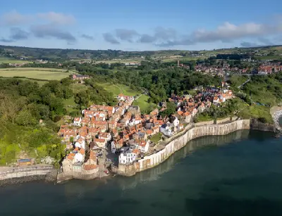 Aerial photograph of Robin Hood's Bay in North Yorkshire, showing red-roofed cottages beside the harbour, coastal seawalls and green hills inland.