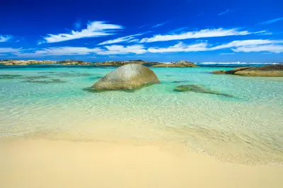 Clear, shallow ocean water gently laps onto the pristine sandy shoreline under a vibrant blue sky with wispy clouds.
