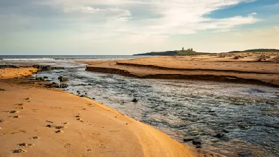 Embleton Burn flowing across the sandy beach into the North Sea at Embleton Bay, with Dunstanburgh Castle visible on the Northumberland coastline.
