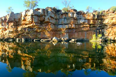 Still water reflects layered sandstone cliffs and scattered vegetation at Bell Gorge in the Kimberley.