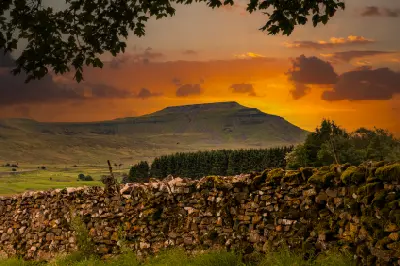 Composite image of Ingleborough mountain beneath an orange sunset sky, framed by trees and a dry-stone wall in the Yorkshire Dales.