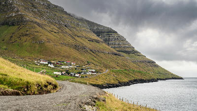 Empty gravel road along the fjord approaching Kunoy village at the base of tall terraced basalt mountains on Kunoy Island, Faroe Islands.