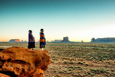 Two people standing on a sandstone outcrop overlooking the desert landscape of Monument Valley in the Southwest United States.