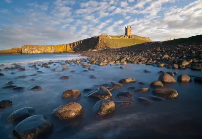 Dunstanburgh Castle tower on a grassy coastal headland above rocky Northumberland cliffs, with smooth long-exposure seawater flowing around foreground boulders in warm afternoon light.