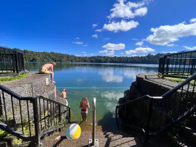 Visitors swimming and entering Lake Eacham from steps with metal railings, overlooking calm blue water and a forested shoreline in Queensland, Australia.