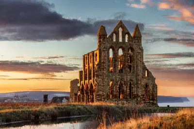 Whitby Abbey ruins illuminated by golden evening sunlight, showing Gothic arches and towers above the North Yorkshire coastline, England.
