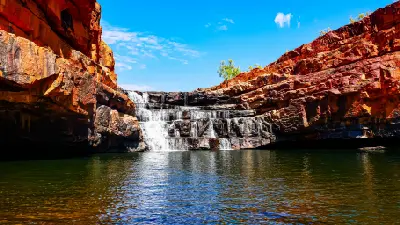 Bell Gorge waterfall cascades over dark rock ledges into a broad pool surrounded by vivid red Kimberley cliffs.