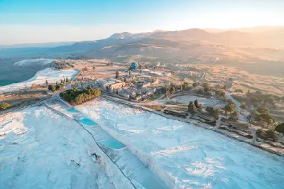 Aerial view of white travertine terraces and turquoise pools at Pamukkale beside the ancient ruins of Hierapolis, with a hot-air balloon and mountains in the distance.