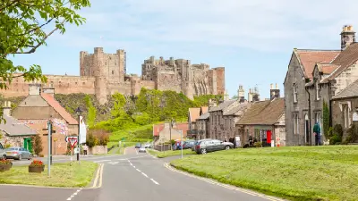View along a village street in Bamburgh, Northumberland, with traditional stone cottages and Bamburgh Castle standing prominently on the hill behind.